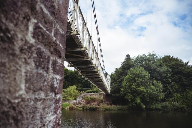 Low angle view of an old bridge spanning over a calm river with lush green trees and a partly cloudy sky in the background. Ideal for use in travel brochures, architectural studies, nature magazines, or websites focusing on historic structures and scenic landscapes.