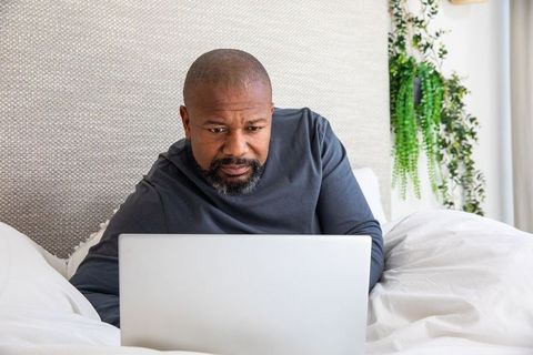 Mature african american man reclining in bed using laptop for remote work and relaxation