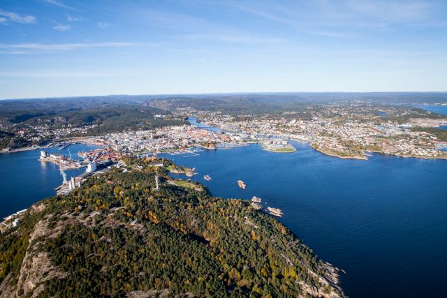Aerial view of a picturesque coastal town surrounded by blue waters and green landscapes under a clear sky. Harbor with numerous boats and an urban cityscape in the distance. Perfect for travel brochures, tourism websites, promotional materials, and city guides.