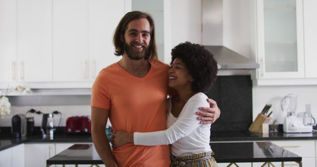 A cheerful interracial couple is embracing and smiling in a modern kitchen. Suggested uses include illustrating content related to relationships, family life, home interiors, lifestyle blogs, domestic happiness, and marketing materials for home appliances.