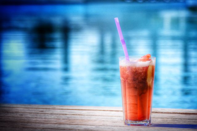 Glass of iced tea with purple straw sitting on wooden deck near swimming pool. Perfect for promoting summer vacations, resort advertisements, poolside relaxation, tropical settings, beverages, refreshing drinks, and leisure activities.