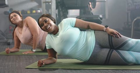 Plus-size women doing side-plank on yoga mats in gym smiling and building core strength