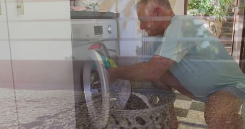 Senior man loading front-load washer with woven basket, kneeling by glass door reflections