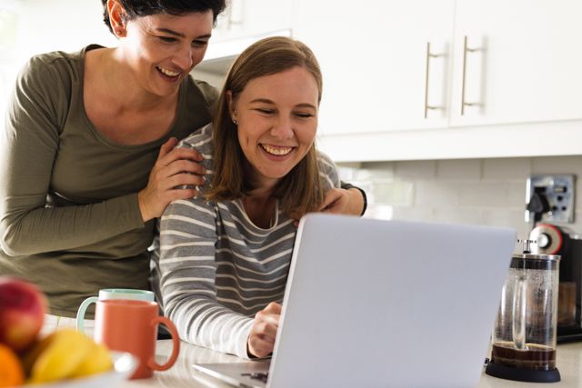 Happy caucasian lesbian woman with hands on shoulders of girlfriend using laptop on kitchen island. Laughing, wireless technology, unaltered, love, togetherness, homosexual, lifestyle and home.