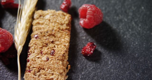 The close-up view showcases a granola bar with its textured surface, accompanied by fresh raspberries and a sprig of wheat on a sleek slate background. This image is ideal for use in health and nutrition blogs, promoting healthy eating, and advertising natural food products.