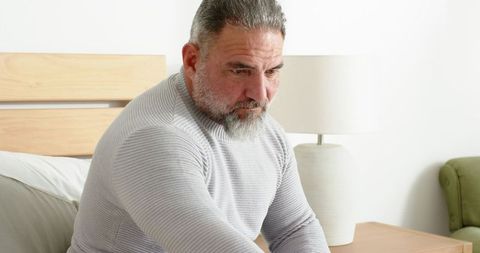 Mature man sitting on bed contemplating in bright minimalist bedroom in light gray sweater