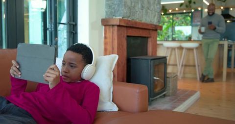 African american boy relaxing with headphones and tablet on couch while father holding mug