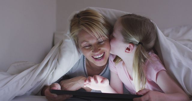 Happy caucasian mother with daughter kissing her and using tablet together under duvet in bed. Motherhood, childhood, love, togetherness and domestic life.