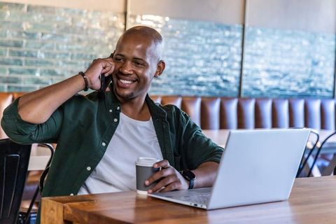 Smiling african american man working remotely in cozy cafe with laptop and coffee