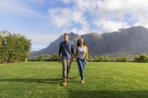 Young couple walking hand-in-hand on lush lawn with mountain backdrop and blue sky