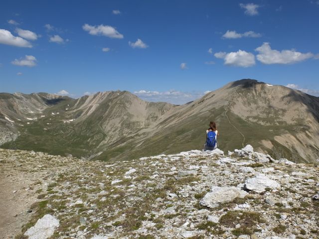 Solo hiker sitting on rocky mountain peak, admiring expansive view of distant mountain range and clear blue sky. Represents themes of adventure, travel, solitude, and natural beauty. Ideal for use in travel blogs, outdoor adventure promotions, environmental campaigns, or motivational material highlighting exploration and perseverance.