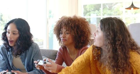 Multicultural women playing video games on sofa, laughing and competing in living room