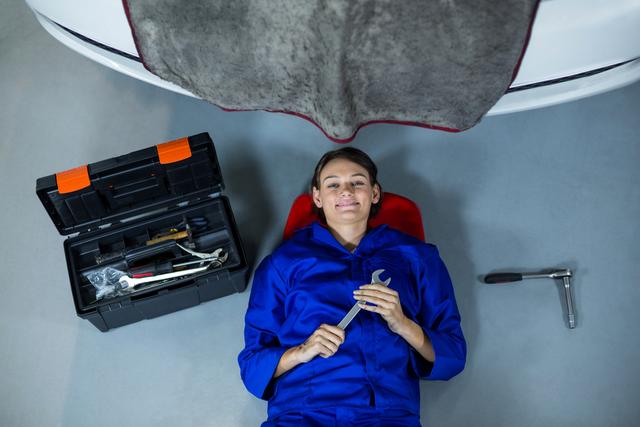 Female mechanic lying under a car, smiling while holding a wrench in an automotive repair garage. She is wearing blue overalls, with a toolbox and other tools beside her. This can be used for promoting auto repair services, advertising mechanical training, or illustrating articles about women in technical professions.