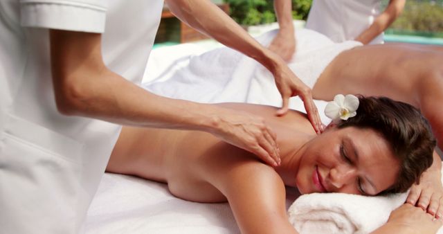 Woman receiving relaxing massage in outdoor spa, lying face down with flower tucked in hair, embodying tranquility and wellness. Ideal for use in promotions for spas, health and wellness centers, skin care products, and travel destinations focusing on relaxation and self-care.