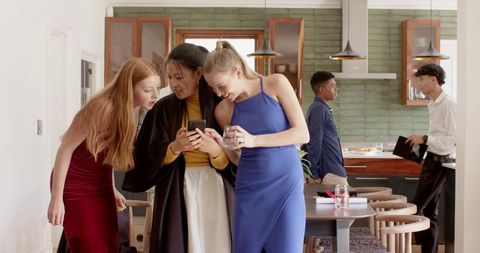 Friends leaning in sharing smartphone at modern kitchen gathering with guests mingling