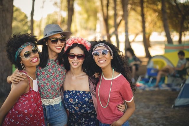Group of female friends smiling and enjoying their time together at a campsite. They are wearing casual summer outfits and sunglasses, with trees and tents in the background. Perfect for use in advertisements, travel blogs, social media posts, and articles about friendship, outdoor activities, and summer adventures.