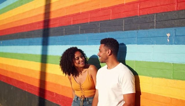 Young couple leaning and laughing by vibrant rainbow striped brick wall on sunny street