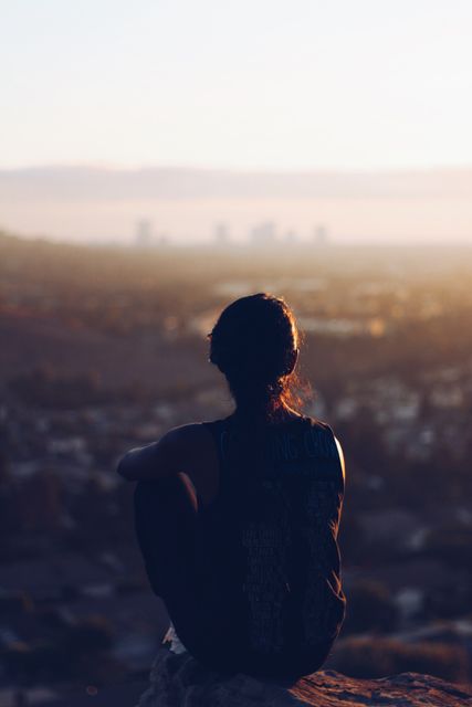 Woman sitting on a hilltop, gazing at a distant city bathed in warm sunlight. Ideal for themes of meditation, contemplation, peacefulness, and connecting with nature.
