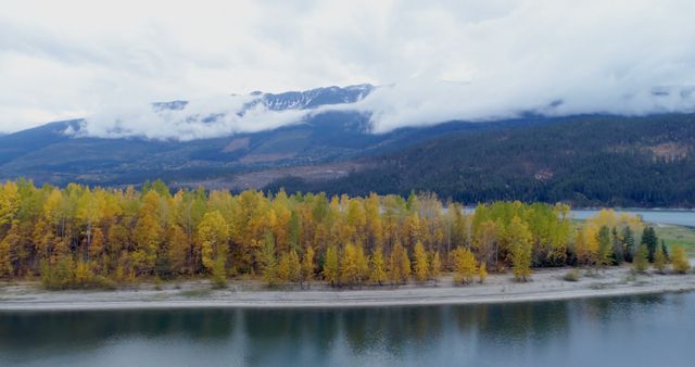 Photo showcasing beautiful autumn landscape with vibrantly colored trees along lake shore. Snow-capped mountains and clouds create serene backdrop. Ideal for use in nature-related articles, travel blogs, and advertisements promoting outdoor destinations or seasonal activities.