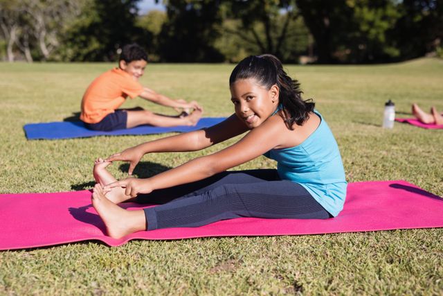 Children practicing yoga outdoors on exercise mats in a park. The girl in the foreground is stretching and touching her toes, while another child is seen in the background. This image can be used for promoting children's fitness, outdoor activities, healthy lifestyles, and wellness programs.