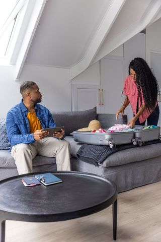 Black couple packing suitcase on gray sectional sofa in cozy attic loft with skylight