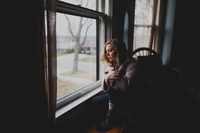 Young woman sitting by window at home, looking outside with a contemplative expression. Perfect for illustrating themes like introspection, tranquility, loneliness, mental health, and peaceful home environments. Suitable for use in blog posts, articles on well-being, home decor ads, and social media content.