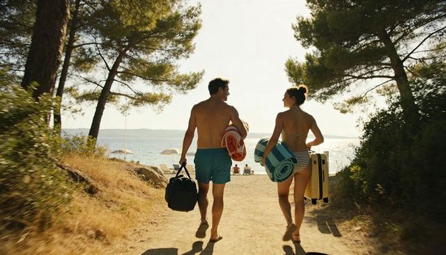 Couple walking to beach carrying towels and luggage on sunlit coastal path