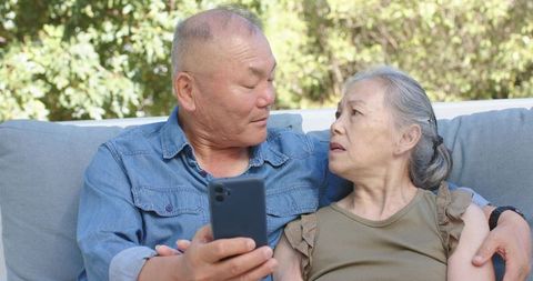 Senior asian couple sharing smartphone moment on garden sofa showing emotional connection