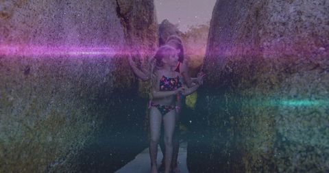 Girls wading through narrow tidal channel between algae-covered boulders with sun flare