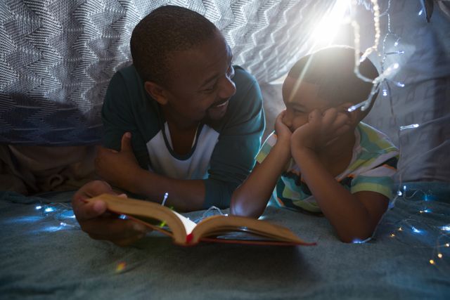 Father and son enjoying quality time reading a book together in a cozy homemade fort with string lights. Perfect for themes related to family bonding, parenting, childhood memories, and indoor activities.