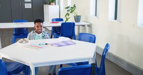 Young African boy sitting at white hexagonal table with blue chairs in bright classroom