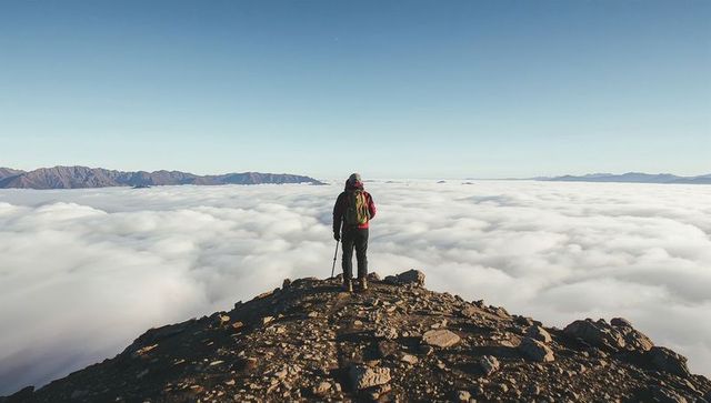 Standing hiker gazing over sea of clouds from rocky summit, backpacking high alpine vista