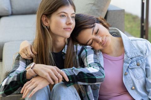 Young women cuddling on patio sofa, leaning head on shoulder, casual denim and plaid
