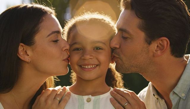 Parents kissing daughter on cheeks in golden hour backyard embracing family warmth affection