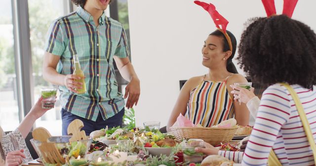 Young multiracial group of friends enjoying a festive meal together indoors. Some wear playful reindeer antlers, adding a fun, spontaneous touch. Ideal for holiday promotions, articles on social gatherings, and lifestyle blogs focused on friendship and celebrations.