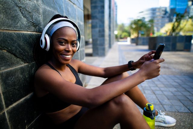 This image shows a smiling African American woman wearing headphones and using a smartphone while resting outdoors. Ideal for promoting healthy lifestyles, fitness apps, workout gear, and urban exercise routines.