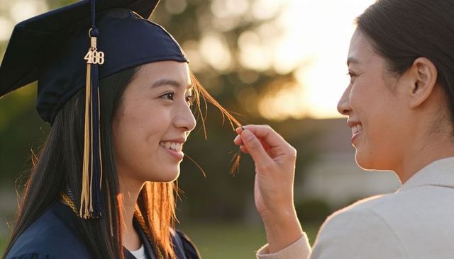Mother tucking hair of smiling graduate in navy cap and gown at golden hour celebration