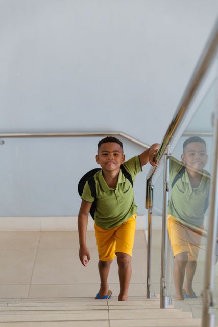 Young African American boy climbing stairs in a school building, holding onto the railing. He is wearing a green shirt and yellow shorts, with a backpack on his shoulder. This image can be used for educational materials, school promotions, back-to-school campaigns, and articles on childhood education and student life.