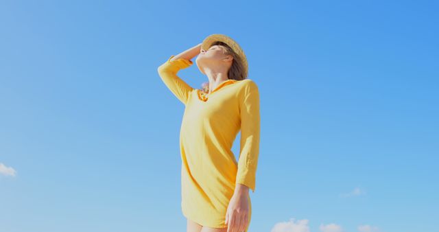 A young Caucasian woman in a yellow dress and straw hat looks up towards the sky, with copy space. Her relaxed pose and the clear blue sky suggest a serene, sunny day ideal for leisure or vacation.