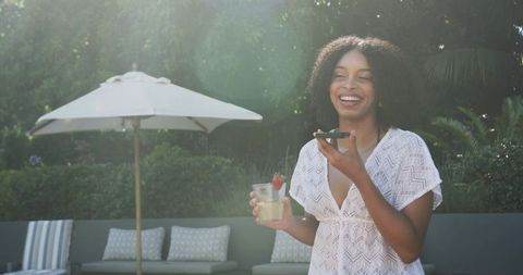 Smiling woman enjoying drink and talking on phone at sunny poolside patio