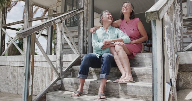 Happy senior caucasian couple embracing on stairs of house on beach, copy space. Senior lifestyle, nature, relaxation, vacation, summer and leisure, unaltered.