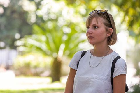 Young woman standing in sunlit park wearing white tee, backpack and sunglasses on head