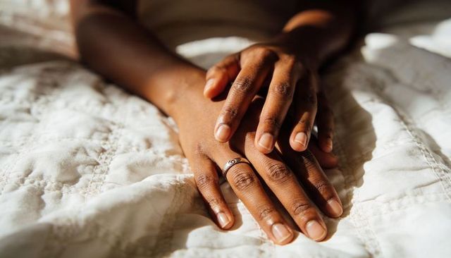 Resting sunlit hands wearing ring on white quilted bedding, close-up of fingers and texture