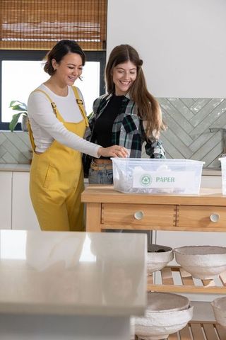 Two women sorting paper for recycling in modern kitchen, smiling eco friendly teamwork