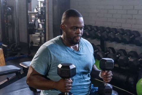 African american man lifting hex dumbbells seated on bench in gym for strength training