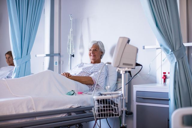 Senior female patient smiling and relaxing on a hospital bed in a ward. She is wearing a hospital gown and has an intravenous drip attached. The scene suggests a positive recovery and professional medical care. This image can be used for healthcare advertisements, medical articles, patient care brochures, and hospital websites.
