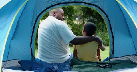 African-american father embracing son while camping in backyard tent