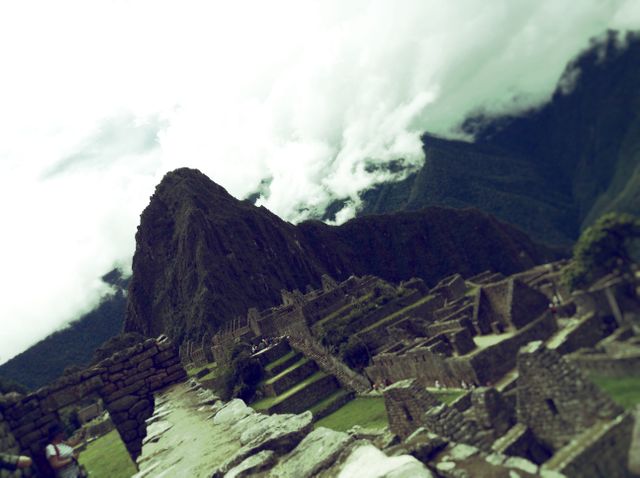 The photo captures a scenic view of Machu Picchu ruins with a mountain covered partially by clouds in the background. Stone structures and terraced hills add depth to the mesmerizing historical landscape. Ideal for use in travel blogs, historical articles, educational content, and posters promoting tourism in Peru.