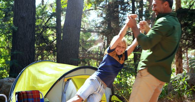 A middle-aged man is playfully swinging a young girl by her arms outside a tent in a forest setting, with copy space. Their joyful interaction suggests a family camping trip and the importance of quality time in nature.