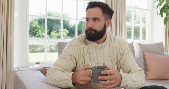A man with a beard sits in a cozy living room, holding a coffee mug and wearing a knitted sweater. Sunlight streams through large windows in the background. This image is perfect for use in advertisements for coffee brands, home decor, leisure activities, and lifestyle blogs.